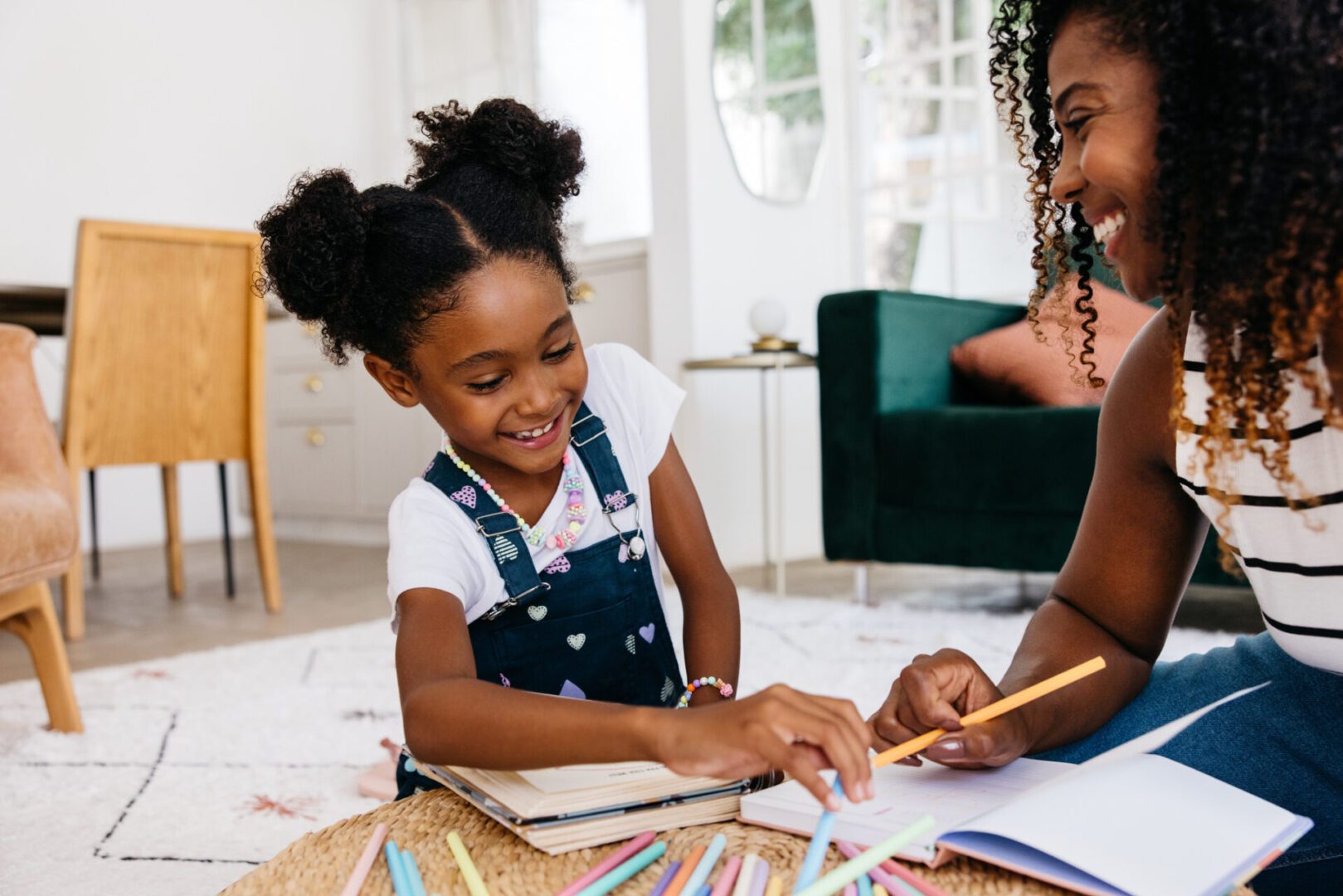 A young girl and her mother laugh and smile while drawing together with colored pencils in the living room at home